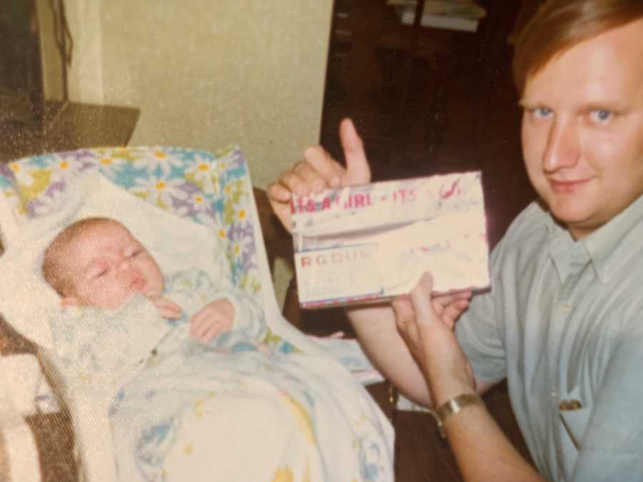 Man holds up announcement for baby girl beside adopted daughter who lays in cradle