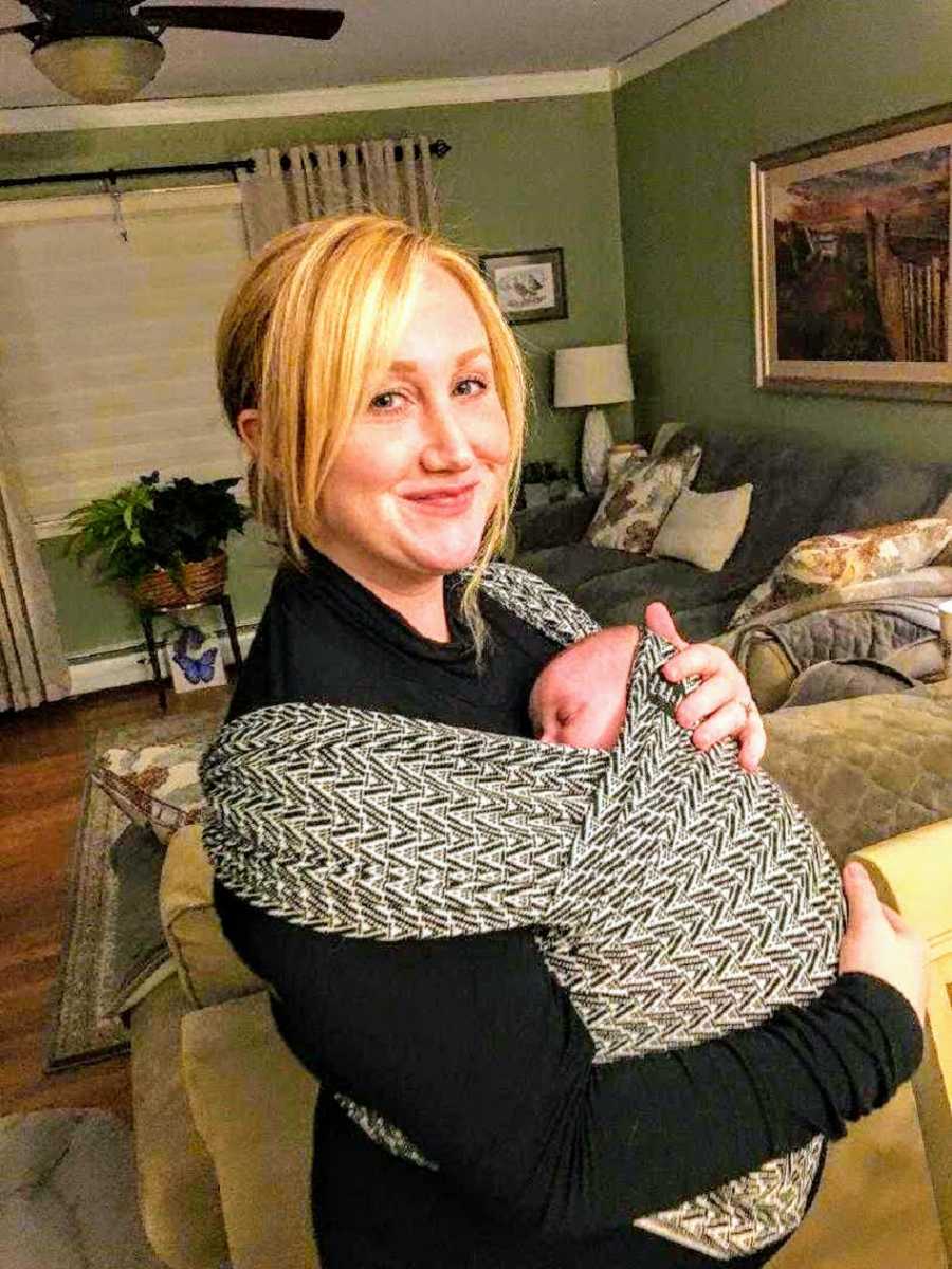 Woman who lost her sister stands smiling in home with baby swaddled to her chest