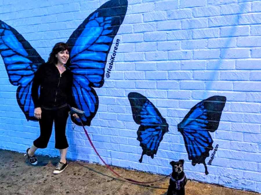 Woman stands in front of butterfly painting on wall while she holds leash that her dog is on