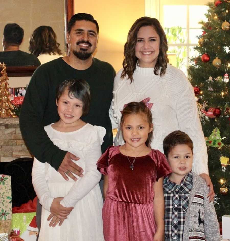 Woman stands smiling with her two kids beside boyfriend and her daughter in front of Christmas tree