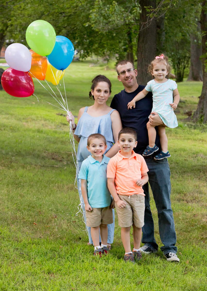 Husband stands outside holding their daughter beside his pregnant wife who holds bouquet of balloons with their two sons in front of them