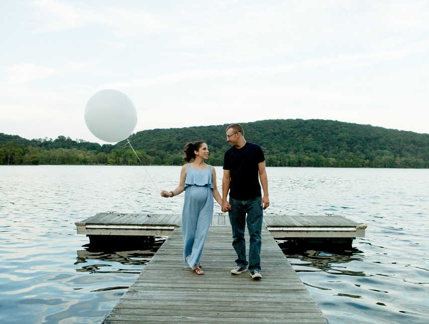 Pregnant woman walks on dock while holding balloon in one hand and husband's hand in the other