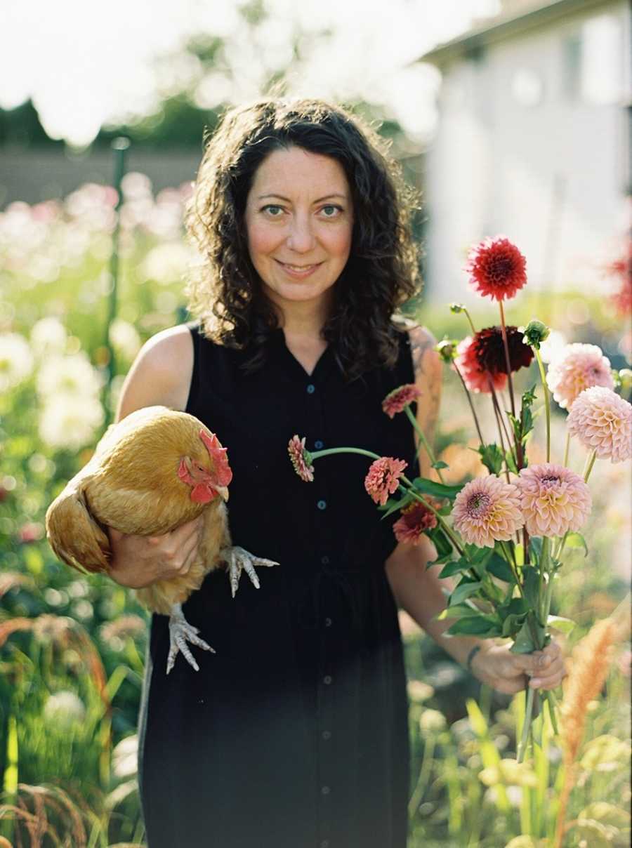 Woman smiles as she stands outside holding flowers in one hand and chicken in other