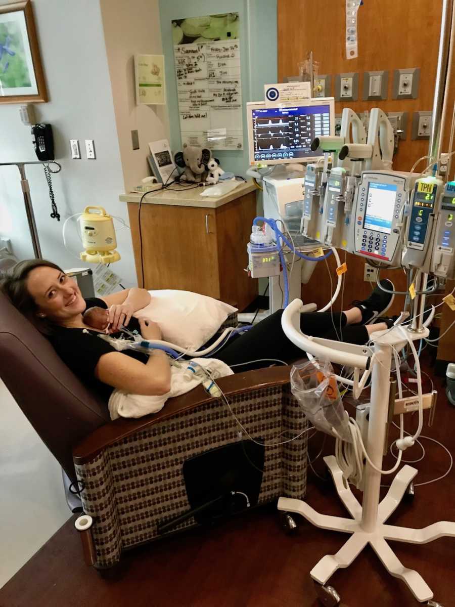 Mother smiles in chair as she holds newborn in NICU in her arms
