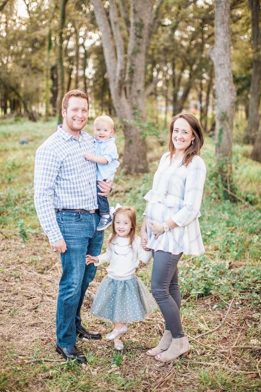 Pregnant woman stands outside smiling in wooded area with daughter and husband who holds son