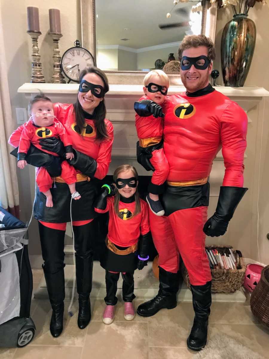 Husband and wife stand in home with their three children all dressed as the family from the Incredibles