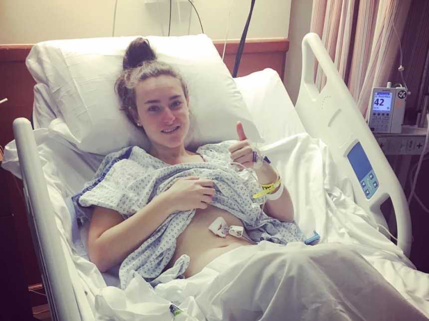 Young woman with MALS lays in hospital bed with thumb up and hospital gown raised to show her feeding tubes