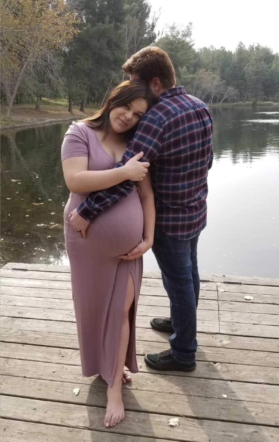 Pregnant woman stands on dock beside husband as they hold onto her stomach