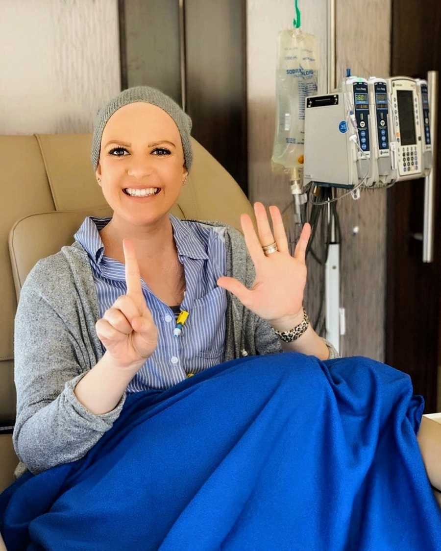 Woman smiles as she sits in hospital chair going through chemo holding up number six
