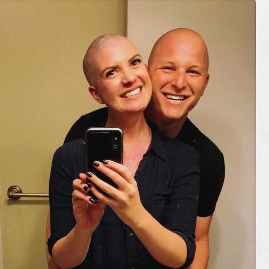 Woman who is finished with chemo smiles as she takes mirror selfie with husband in background