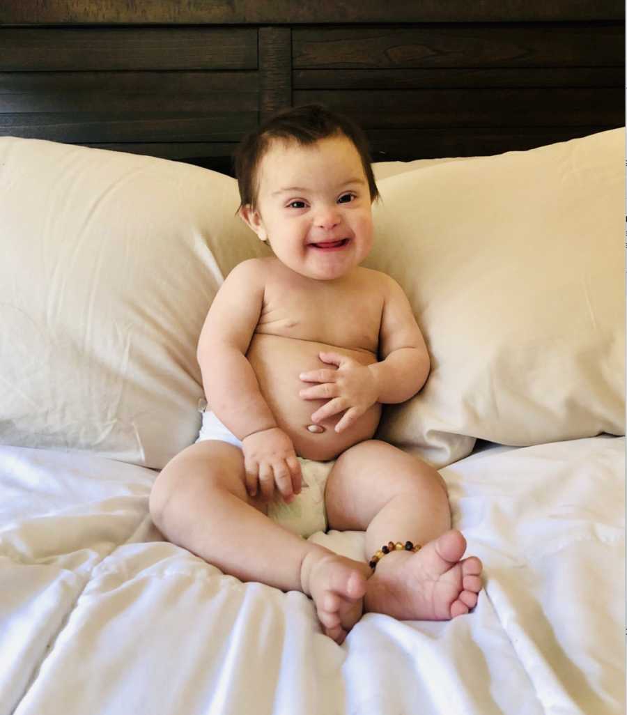 Baby girl with down syndrome sits in bed wearing just a diaper smiling