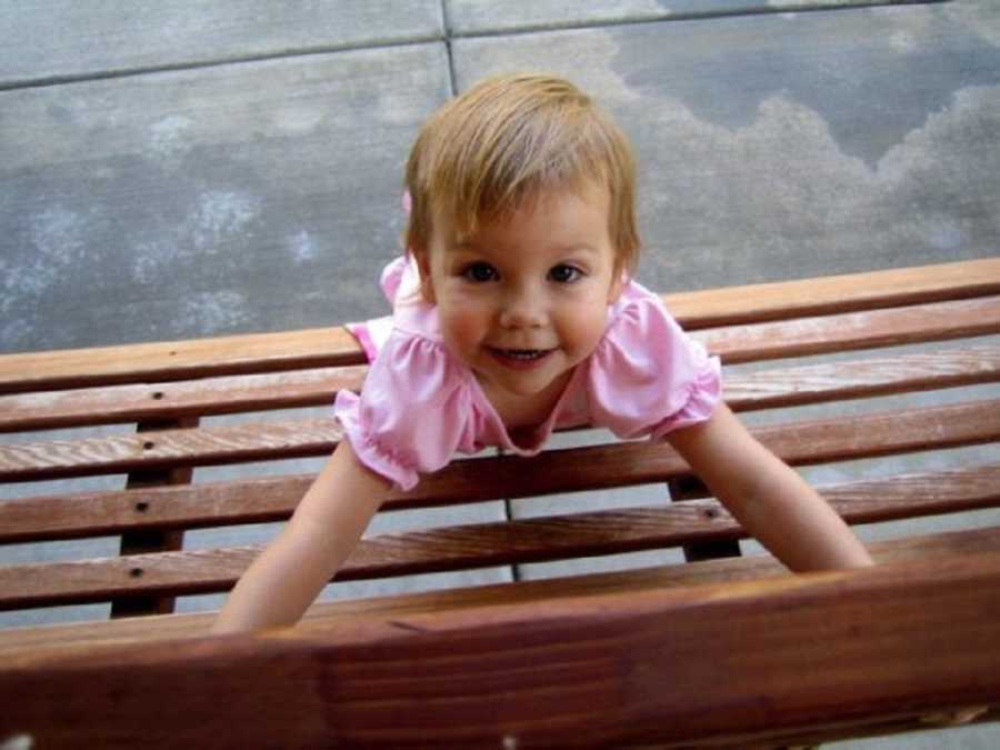 Little girl smiles as she leans over wooden bench outside 