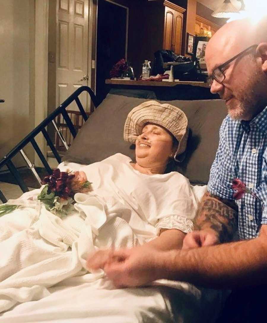 Woman with terminal cancer smiles in bed in home holding hands with man who she just married
