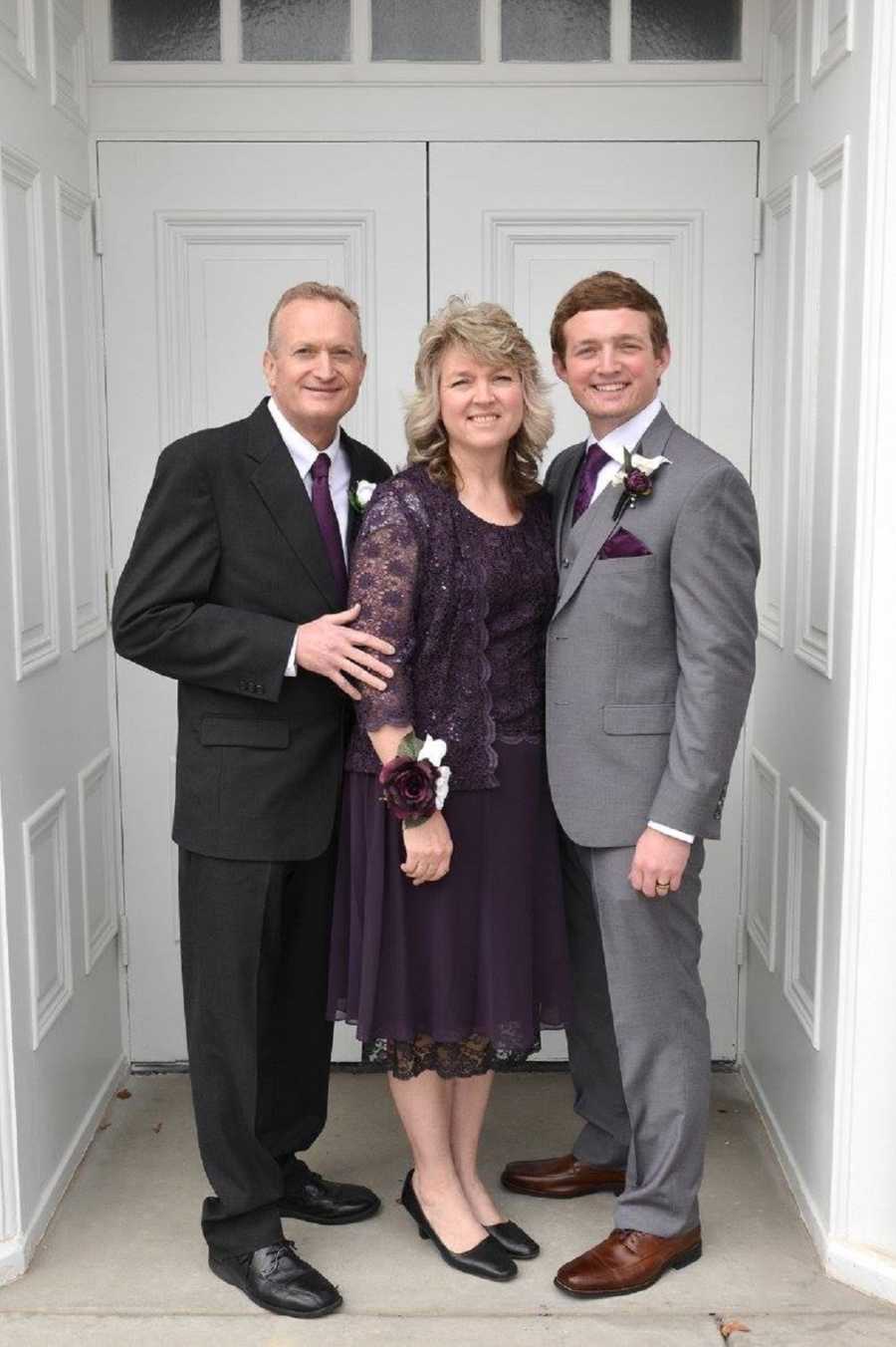 Young man stands outside of church in suit beside mother and father