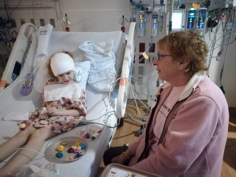 Little girl with brain cancer sits in hospital bed with bandage over her head as grandmother sits at her side