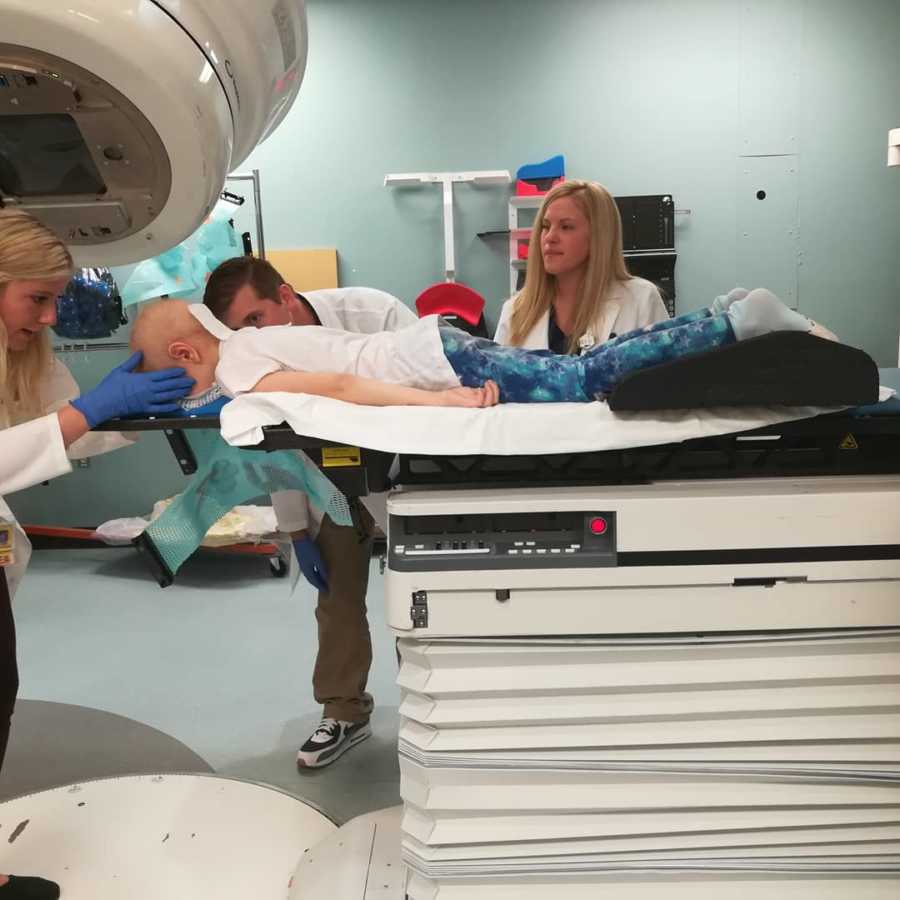 Young girl with brain cancer lays on her stomach on hospital bed awaiting radiation with her team at her side