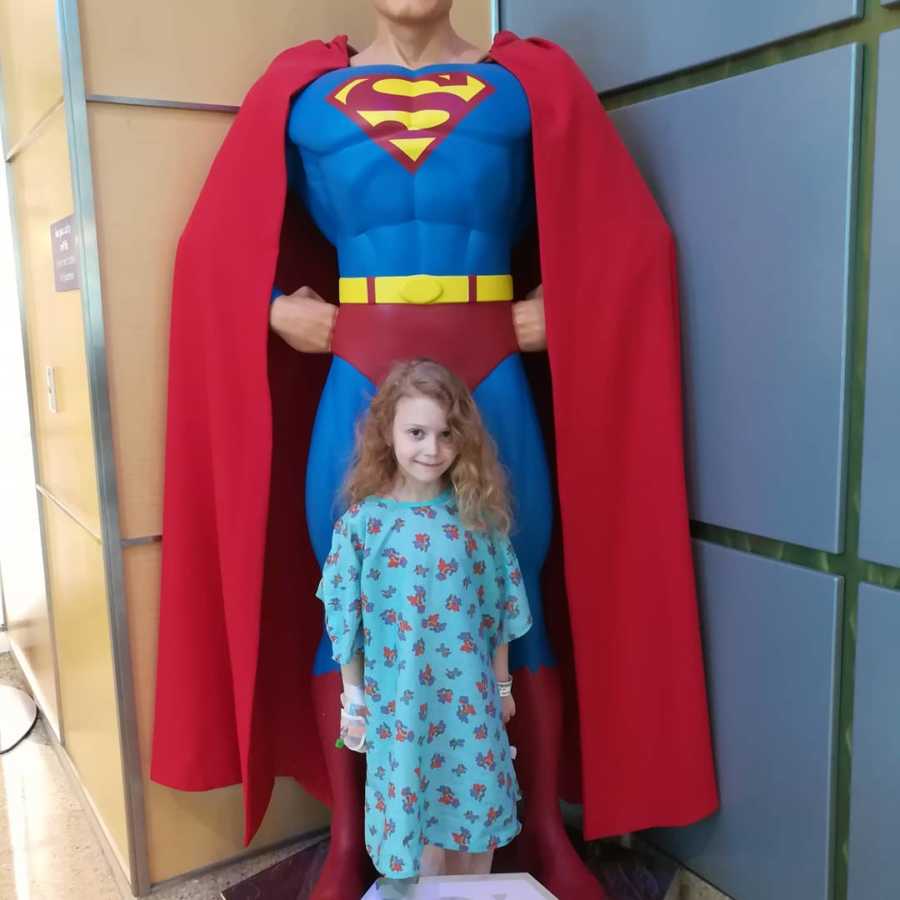 Little girl with brain cancer stands in hospital gown in front of Superman statue