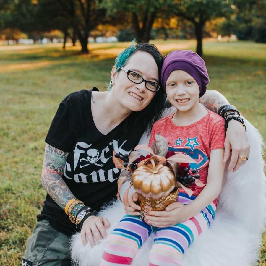 Little girl with cancer sits in white fuzzy chair outside as mother crouches beside her with arm around her