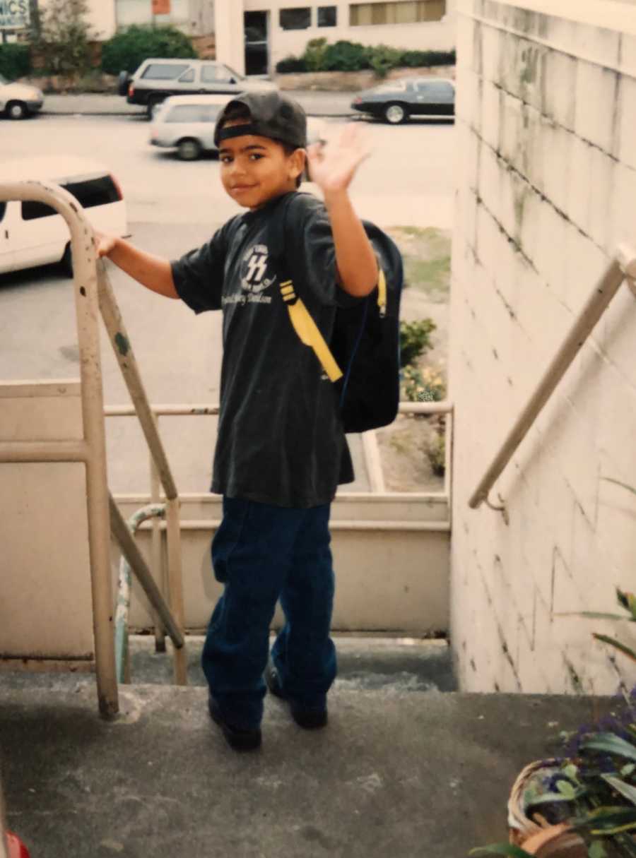 Little boy with backpack stands at top of steps looking over his shoulder waving