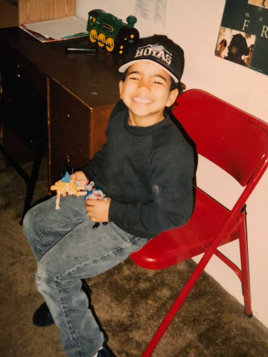 Little boy who grows up to be drug addict sits in red fold up chair smiling as he holds toys