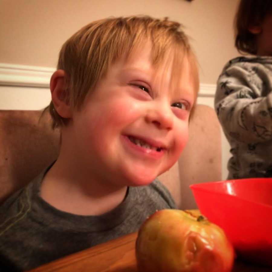 Little boy with down syndrome sits smiling at table with apple in front of him