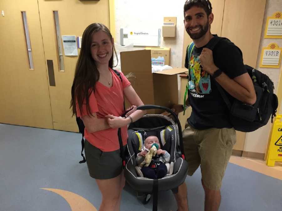 Husband and wife smile as they stand in hospital hallway with baby in carseat