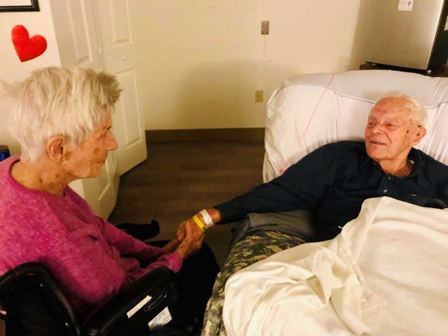 Elderly woman with dementia sits in wheel chair holding husband's hand who lays in hospital bed