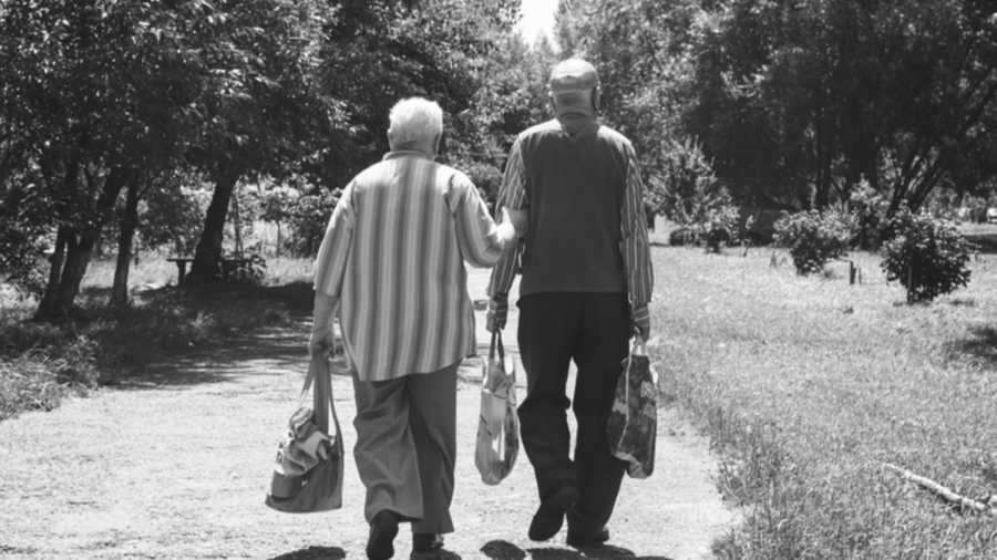 Elderly woman walks outside holding bag and arm of husband who holds two bags