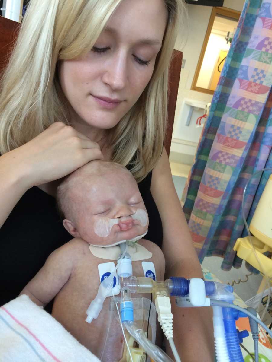 Mother sits in NICU looking down at newborn in her lap with heart issues and trach