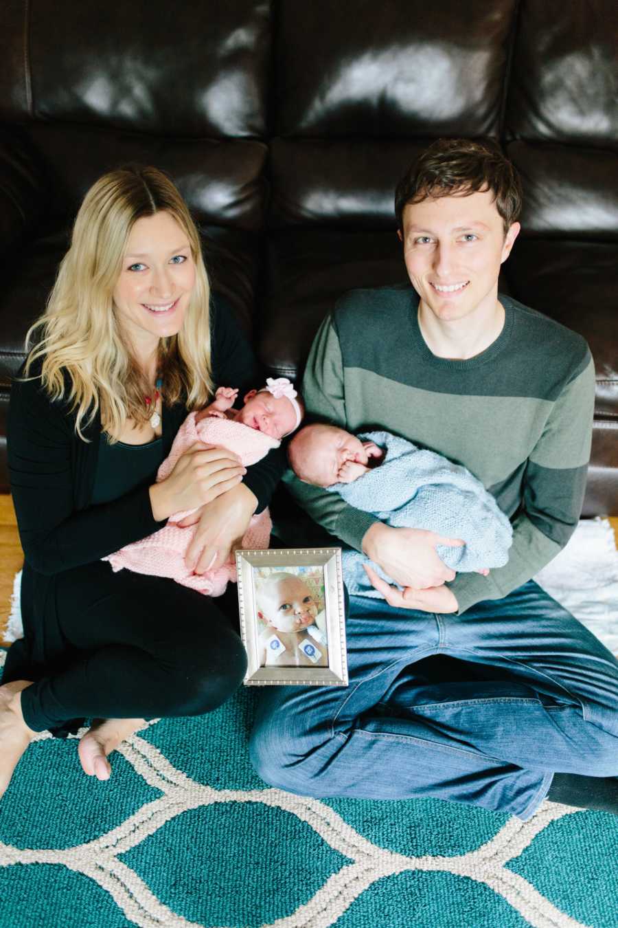 Husband and wife sit on floor of home smiling as they hold their newborn twin with picture of their deceased first baby in front of them