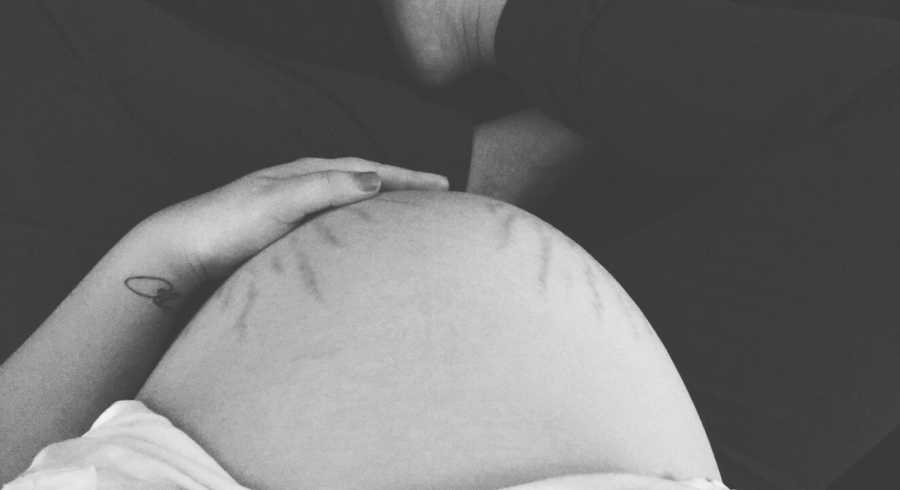 Pregnant woman sits on floor holding hand to stomach with stretch marks on it