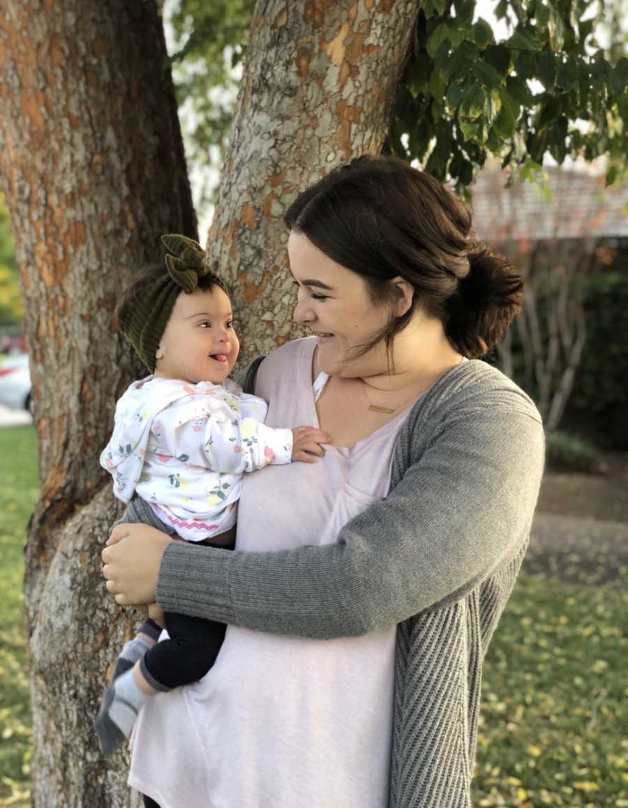 Mother stands outside beside tree smiling at daughter with down syndrome in her arms