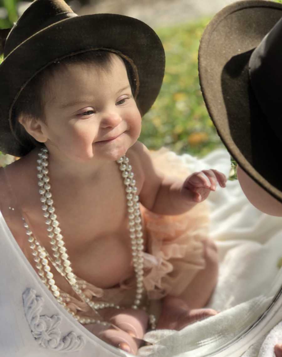 Little girl with down syndrome sits on ground wearing pearl necklace and hat as she smiles in mirror