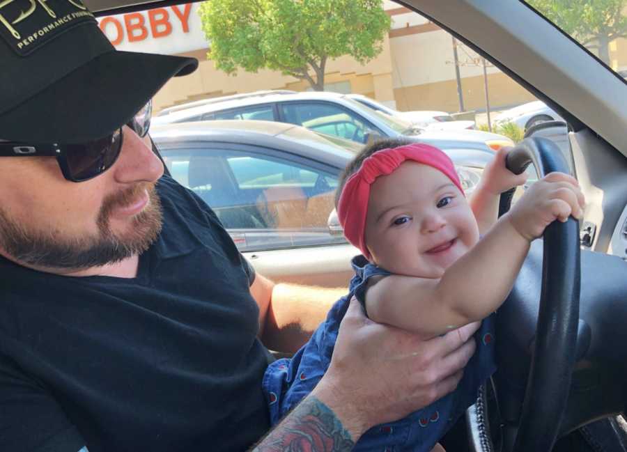 Baby girl with down syndrome sits in fathers lap in car holding onto steering wheel