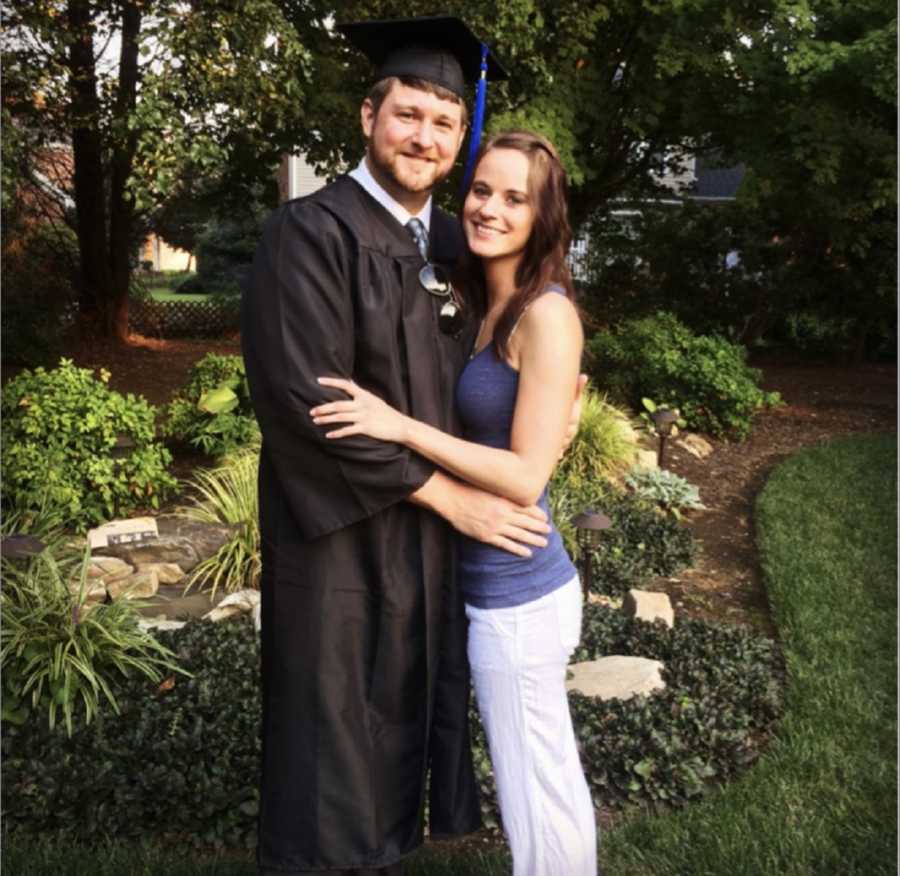 Boyfriend in cap in gown stands outside holding onto girlfriend as they smile