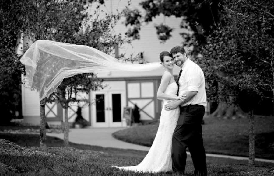 Bride and groom stand outside in each others arms as bride's veil flies in the wind