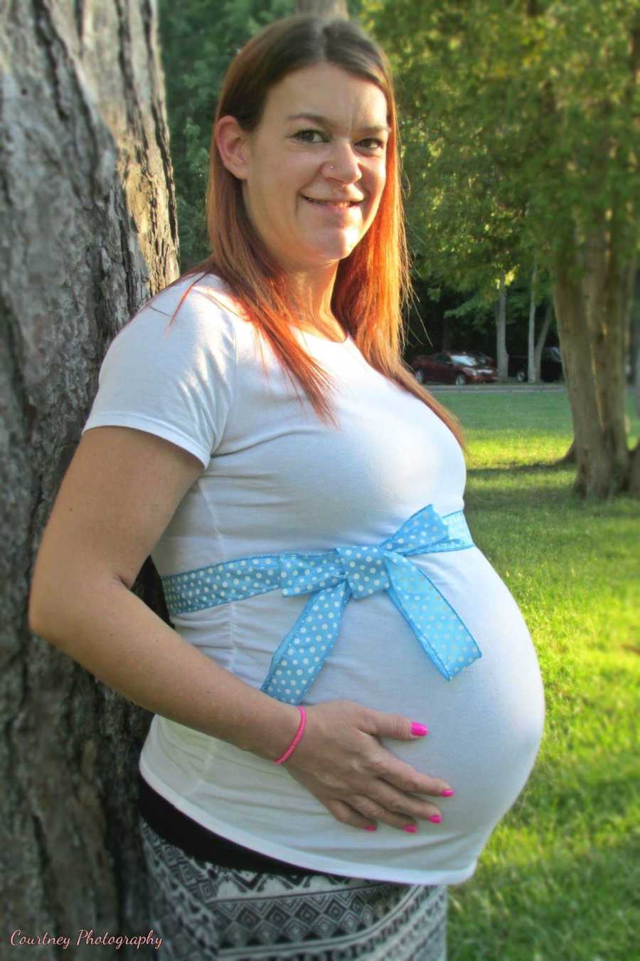Pregnant woman stands outside beside tree with blue bow wrapped around her stomach