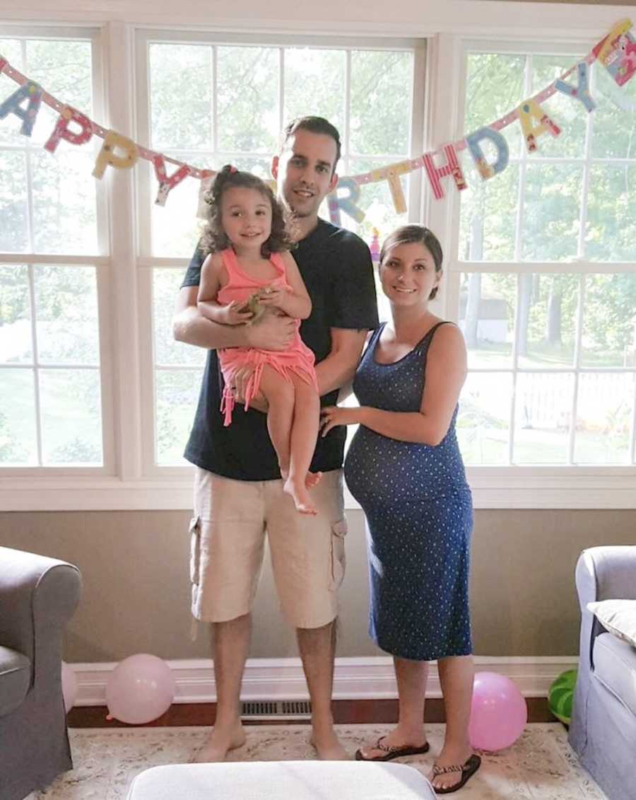 Pregnant woman stands in home beside husband who holds their daughter in front of "Happy Birthday" sign