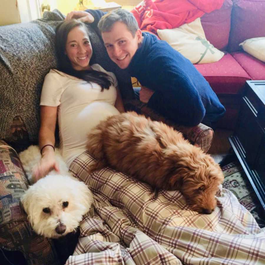 Pregnant woman smiles as she lays on couch with two dogs and her husband kneels beside her