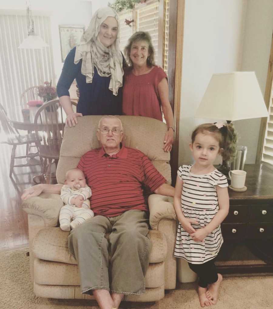 Elderly man sits in chair with baby as young girl stands beside him and wife and daughter behind him