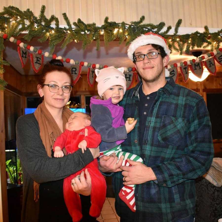 Husband and wife with postpartum depression stand in home decorated for Christmas holding their two children