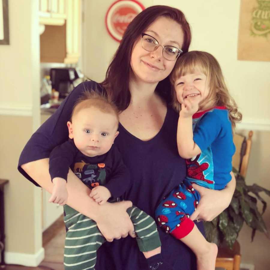 Mother with PPD stands smiling in home while she holds her two young kids