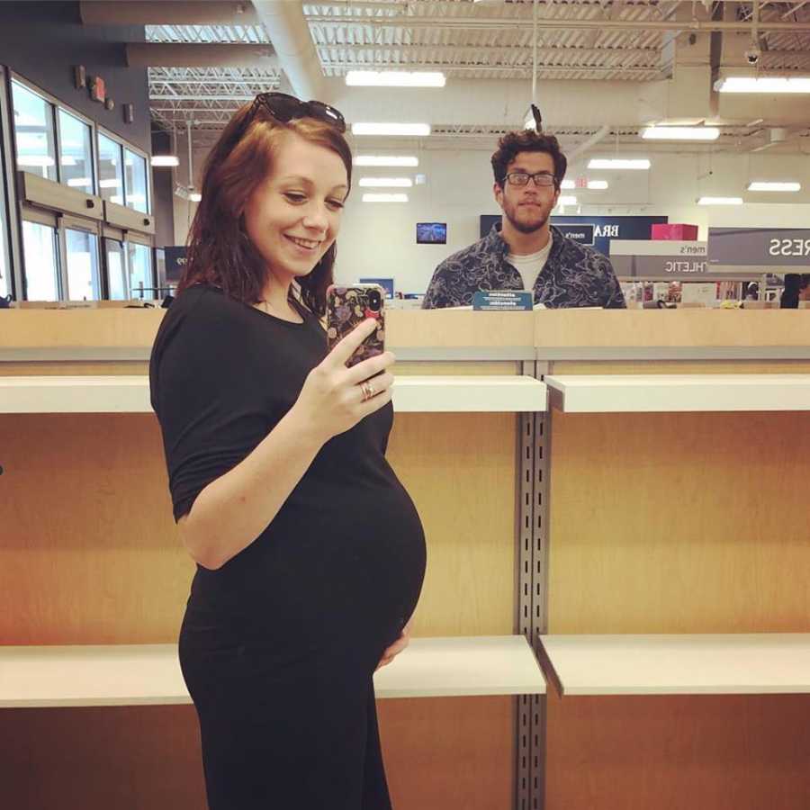 Pregnant woman smiles as she takes mirror selfie in empty store aisle with husband in background