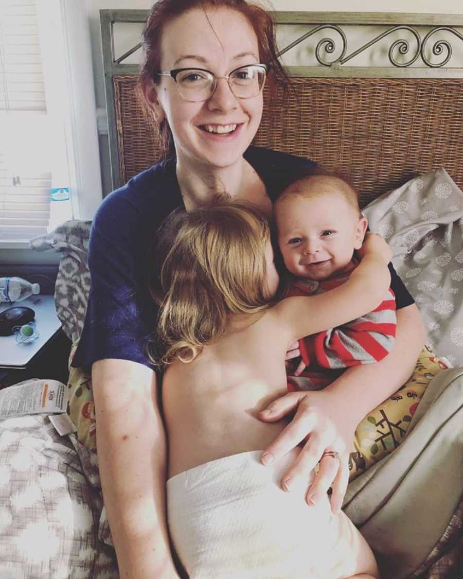Mother who finally loves herself sits smiling in bed with her two kids in her lap