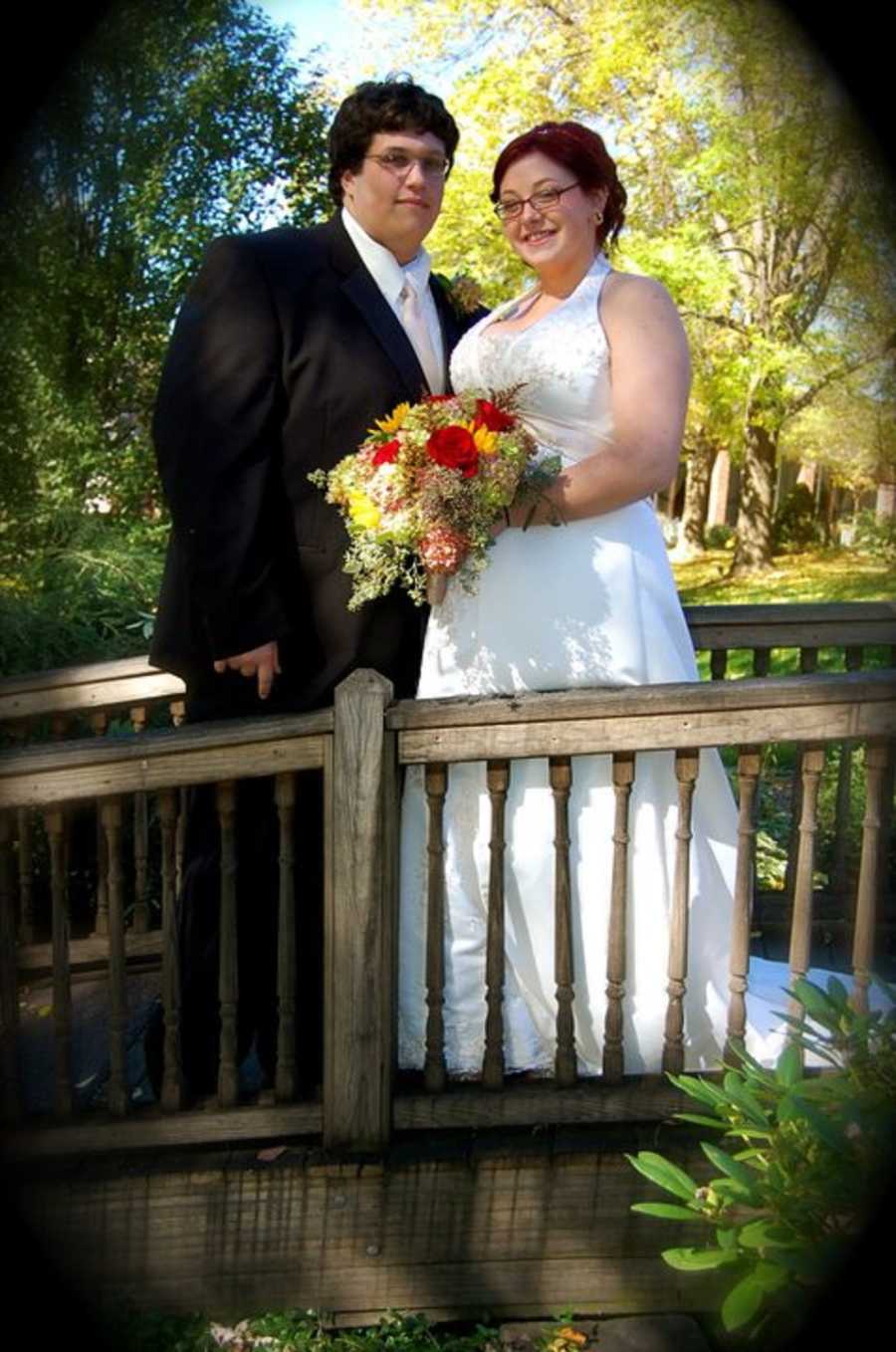 Bride with eating disorder stands on wooden bridge smiling beside groom