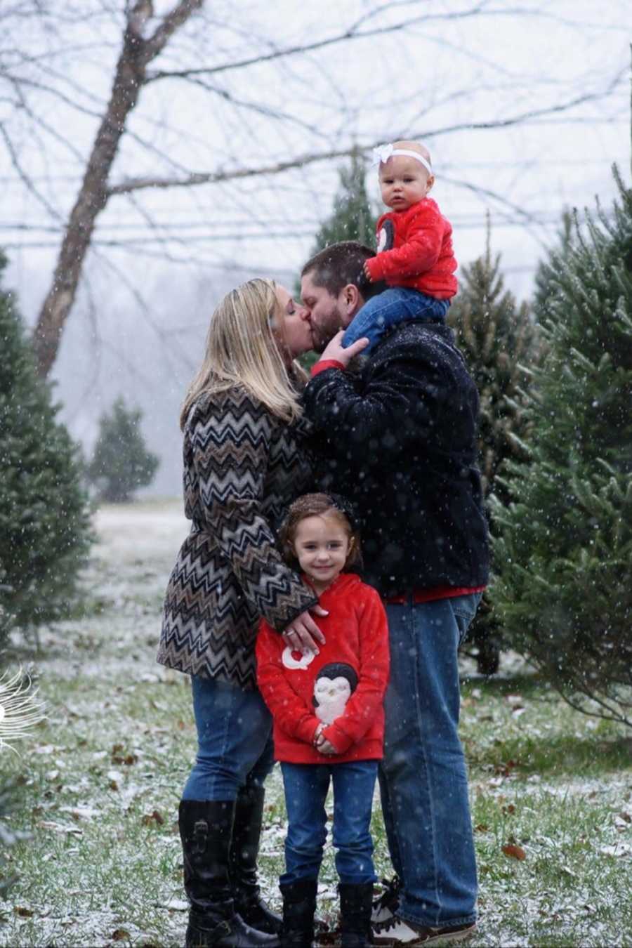 Husband and wife stand outside kissing as it snows while one daughter stands in front of them and other sits on father's shoulders