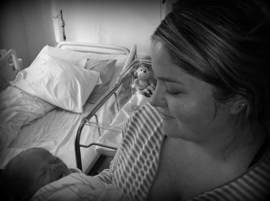 Mother smiles as she stands in hospital room holding her newborn baby who is asleep