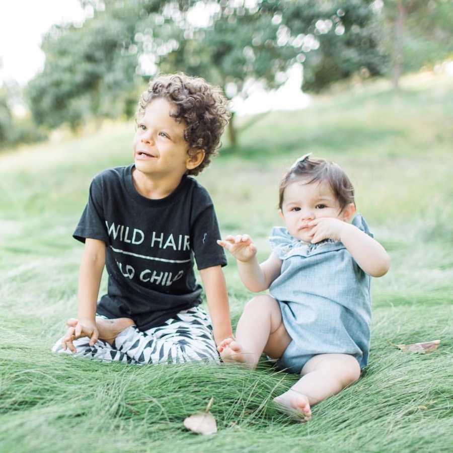 Little boy with Williams Syndrome sits in grass smiling with his little sister sitting beside him