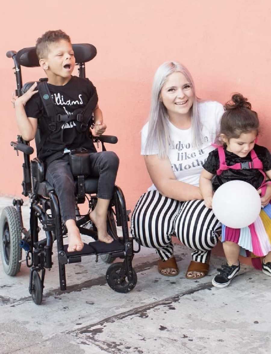 Little boy with Williams Syndrome sits in wheelchair beside his mother who squats down to hug daughter