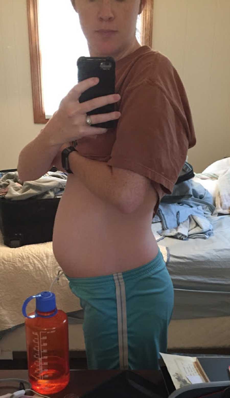 Pregnant woman stands in bedroom with shirt raised in mirror selfie